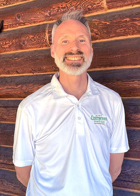 Picture of David Heia, Fair Board Member. White shirt standing in front of log cabin wall.